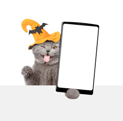 Aufkleber - Winking licking lips kitten wearing hat for halloween holds shows smartphone with white blank screen in it paw above white blank board. Empty space for text. Isolated on white background