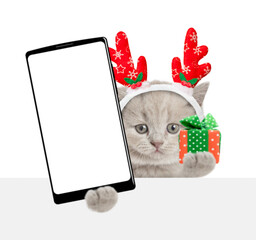 Aufkleber - Cute kitten wearing red christmas hat holding gift box and showing smartphone with white blank screen in it paw looks above blank white banner. Empty space for text. Isolated on white background