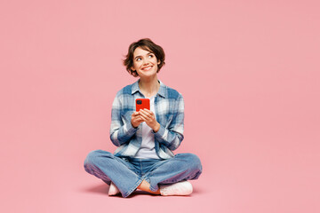 Wall Mural - Full body young woman wear blue shirt white t-shirt casual clothes sits hold in hand use mobile cell phone look aside isolated on plain pastel light pink background studio portrait. Lifestyle concept.