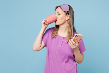 Wall Mural - Calm young woman wears purple pyjamas jam sleep eye mask rest relax at home hold use mobile cell phone drink tea milk isolated on plain pastel light blue cyan background. Good mood night nap concept.