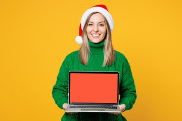 Wall Mural - Merry fun young IT woman wear green sweater Santa hat posing hold use work on blank screen laptop pc computer isolated on plain yellow background. Happy New Year celebration Christmas holiday concept