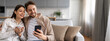 © Prostock-studio - A young couple is sitting comfortably on a couch in their home, sharing a joyful moment as they look at a smartphone while one holds a cup.
