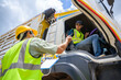 © เลิศลักษณ์ ทิพชัย - A logistics worker in safety gear hands over documents to a truck driver from outside the vehicle, symbolizing coordination and teamwork in transportation and logistics.