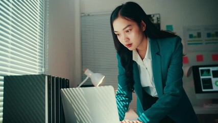 Aufkleber - Young businesswoman in a teal suit leans over a laptop on a desk, focusing intently on analyzing data and managing tasks in a modern office filled with natural light