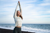Peaceful young woman meditating on seashore at sunrise, sporty girl stretching arms up by the sea and sky, breathing fresh air and doing yoga on beach, morning run, health wellbeing sport activities