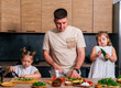 © dvulikaia - Young father cooking with his little daughters. Father teaching kids how to cook.