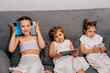 © dvulikaia - Cute little sisters sitting on sofa using modern gadgets to watch videos and play games.