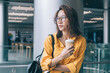 © BullRun - Pensive Chinese hipster girl in classic glasses standing at public place and feeling pondering during free time indoors, attractive contemplative female in optical eyewear for provide eyes protection