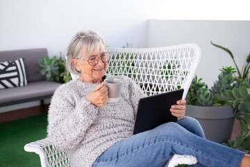 Wall Mural - Attractive senior gray haired woman holding a coffee cup relaxing at the armchair in the balcony at home looking at digital tablet