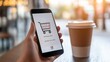 © olga_demina - Hand holding mobile phone displaying shopping cart icon and payment information, suggesting online purchase in cafe setting with takeaway coffee cup