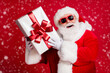 © deagreez - Photo of pensioner old man white beard hold present box ribbon bow-knot curious interested wonder what inside wear santa costume sunglass gloves headwear isolated red color background