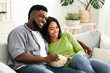 © Prostock-studio - Relaxed african american couple watching tv, sitting on couch with popcorn, enjoying staying at home together