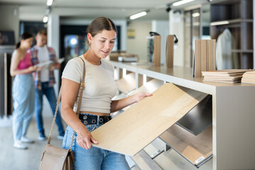 Young, keen female shopper dressed in casual attire exploring various wood-patterned laminate flooring options on sample rack in building hypermarket, choosing perfect design for home renovation