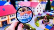 © Sam - A hand holds a magnifying glass over a miniature village, focusing on a small house with a red roof.