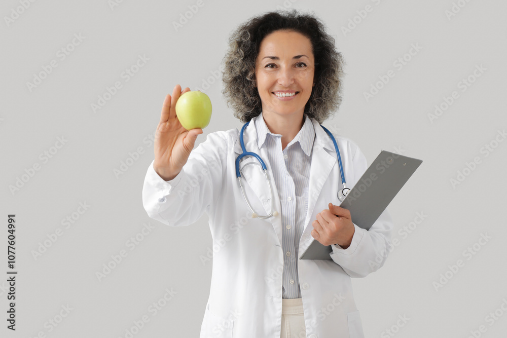 Mature female nutritionist with apple and clipboard on light background