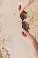 Poster - Hands holding sunglasses on sandy beach with sun drawing