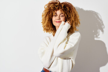 Wall Mural - Portrait of a cheerful woman with curly hair wearing a cozy white sweater, standing against a plain white background, exuding warmth and positivity