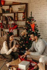 A loving couple is wrapping Christmas presents together at home in a cozy living room, surrounded by ribbons, bows, and wrapping paper. 