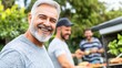 © PPS - A man with a gray beard smiles at the camera while standing in front of two other men who are grilling food outdoors.