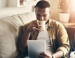 © peopleimages.com - House, coffee and black man on sofa, tablet and reading morning news, relax and internet. African person, apartment and guy on couch, technology or herbal tea with digital app for ebook or connection