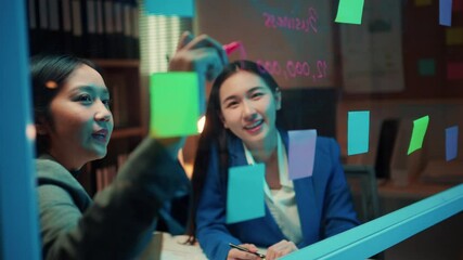 Poster - Two young businesswomen engage in a strategy discussion, utilizing colorful sticky notes on a glass wall in their office during the night, fostering collaboration and creativity