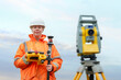 © Iryna - Woamn surveyor in orange jacket using equipment at construction site during cloudy day