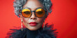 © julijadmi - Confident senior woman with gray hair wearing bold sunglasses and black feathers against a red background.