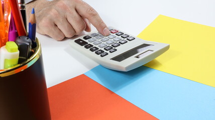 Man's hand typing on calculator Desk with colored papers and stationery out of focus