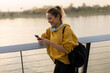 © BGStock72 - Young woman enjoying sunset by the river while texting on her smartphone, wearing a cozy yellow hoodie and headphones