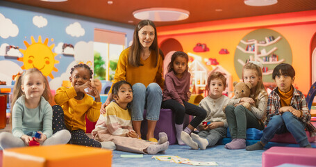 Wall Mural - Portrait of Happy Diverse Children Posing Together with a Female Preschool Teacher in a Modern Kindergarten. Educator and Beautiful Little Kids Looking at Camera, Smiling, Waving Hands