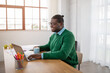 © Prostock-studio - A black guy wearing a green sweater and glasses is working diligently on a laptop at a sunny desk. Nearby, there is a mug and some colorful pens, creating a vibrant workspace.