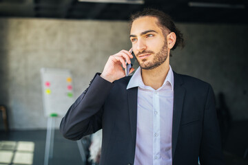 Aufkleber - A young man dressed in a dark suit speaks on his phone while standing in a contemporary office. The environment features bright lighting and a whiteboard in the background.