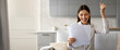 © Prostock-studio - A young woman sits on her couch, holding papers with excitement. She smiles broadly, raising a fist in celebration, indicating a positive outcome. The cozy living space adds to the joyful mood.
