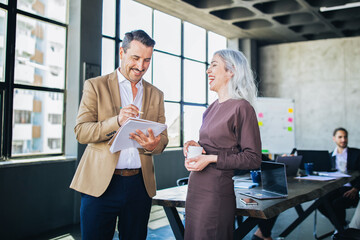 Fototapete - Two colleagues interact in a modern office setting, exchanging ideas and laughter while one takes notes. The lively atmosphere encourages collaboration and creative thinking.