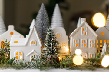 Wall Mural - Christmas still life, miniature winter holiday village. Merry Christmas and Happy Holidays! Christmas white little houses, trees and lights on table with fir branches. Atmospheric magical eve