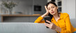 © Prostock-studio - A woman wearing a bright yellow shirt sits on a cozy couch, resting her chin on her hand, looking thoughtfully at her smartphone in a contemporary living space.