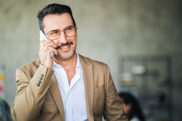 Aufkleber - A man in a tan blazer is smiling while on a phone call in a contemporary office. The background features a blurred workspace, indicating a professional environment.