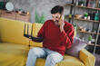 © deagreez - Young man in red sweater frustrated with technology issues while sitting on yellow sofa indoors