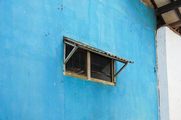 Poster - A single window with a metal awning sits on a blue wall of a building. The window is a simple design with a wooden frame and a metal grid. A small window on a bright blue wall.