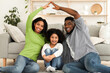 © Prostock-studio - Stay At Home Concept. Happy african parents making symbolic roof of hands above their little daughter's head, sitting together on floor in living room