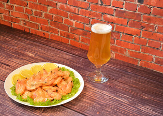 A plate of freshly cooked shrimp on a lettuce leaf with lemon wedges and a tall glass of light beer on a wooden table.