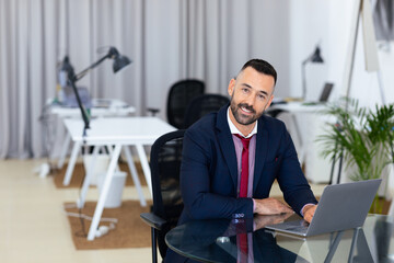 Leinwandbilder - A man in a suit is seated at a glass desk in a contemporary office. He smiles while using a laptop, surrounded by minimalist furniture and indoor plants, creating a productive atmosphere.