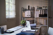 © Pichsakul - An organized office desk with a laptop, documents, and a small potted plant, set against a background of shelves and window blinds