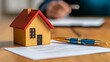 © Pedro - A house model, loan documents, and a pen placed neatly on a table, with an agent and customers talking about mortgage