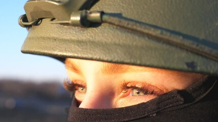 Wall Mural - Close up green eyes of female ukrainian army soldier looking at sunset. Sight of young military woman in helmet and balaclava at countryside. Invasion of territory Ukraine. End of war in Europe