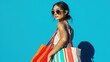 © Thuntaree - Stylish Young Woman Posing with Colorful Beach Bags in Bright Sunglasses Against a Vibrant Blue Background Ready for Summer Fun and Adventures