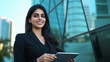 © Maksym - An elegant young Indian businesswoman outdoors at a business complex, looking at her tablet while smiling, with reflections of skyscrapers in the glass behind her.