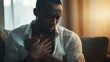 © Lion - Dark-skinned man clutching his chest while seated on a couch, wearing a light shirt, in a softly lit room with a blurred background
