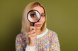 © deagreez - Blonde woman holds magnifying glass to her eye while wearing colorful sweater against olive green background