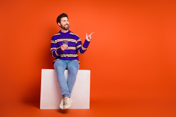 Poster - Full size photo of young male sit white cube look point empty space hold device wear striped violet outfit isolated on orange background